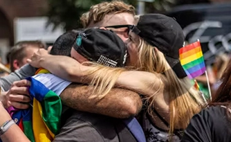 People embracing at a Pride event, with rainbow flags visible in the background.